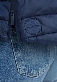 Marineblaue gesteppte Jacke mit Reißverschluss und Gummi-Milestone-Logo-Patch. Getragen über ausgewaschenem Denim, der Textur und Farbkontrast zeigt.