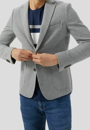 Persoon die een lichtgrijze blazer dichtknopte over een marineblauw overhemd met witte en grijze strepen, draagt een blauwe spijkerbroek, tegen een effen achtergrond.