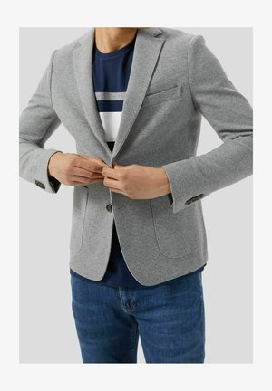 Persoon die een lichtgrijze blazer dichtknopte over een marineblauw overhemd met witte en grijze strepen, draagt een blauwe spijkerbroek, tegen een effen achtergrond.