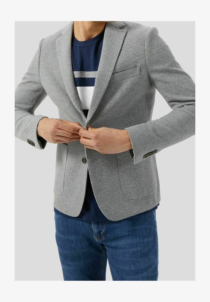 Personne boutonnant un blazer gris clair sur une chemise bleu marine à rayures blanches et grises, portant un jean bleu, sur un fond uni.
