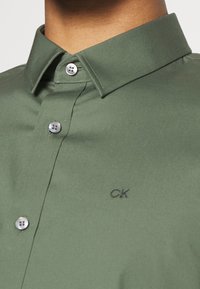Close-up van een persoon die een olijfgroen overhemd met knopen en een puntige kraag draagt, met een klein geborduurd "CK"-logo op de borst.
