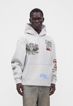 Homme portant un sweat à capuche gris clair avec des patchs graphiques colorés et un jean délavé, debout devant un fond blanc uni.