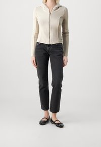 Person klädd i en krämfärgad zip-up hoodie, svarta rakbeniga jeans och svarta spikade Mary Jane-flats, stående mot en enkel ljus bakgrund.