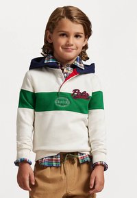 Χαμογελαστός αγόρι φορώντας μια λευκή φούτερ με πράσινη λωρίδα και λογότυπο "Polo", καρώ πουκάμισο από κάτω και καφέ παν trousers σε απλό φόντο.