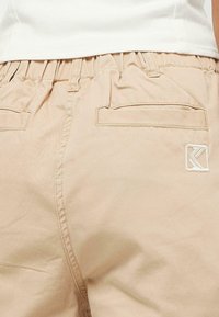 Beige bomullsshorts med elastisk midja, två bakfickor och en broderad logotyp i hörnet. Slät textur, avslappnad design.