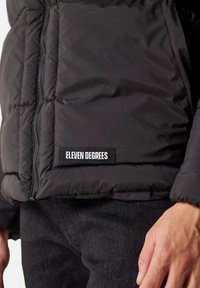 Chaqueta puffer negra con una capa exterior texturizada, cierre de cremallera y un prominente parche de logo "ELEVEN DEGREES" en la parte inferior frontal.