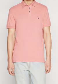 Ljusrosa pikétröja med knappslå, korta ärmar och en liten logotyp på bröstet. Tyget verkar vara bomull eller liknande material.