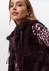 Bordeauxrood faux-fur vest met een gestructureerd, gelaagd ontwerp en een capuchon. Voorzien van een rits aan de voorkant en trekkoordaccenten. Gecombineerd met een gebloemd shirt.