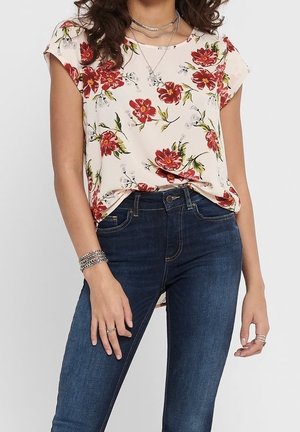 Blouse à manches courtes florale en crème clair, ornée de fleurs rouges et vertes, associée à un jean skinny bleu foncé. Tissu doux, coupe décontractée.