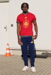 Červený dres španielskeho národného tímu s emblémom, modré tréningové nohavice Adidas a biele tenisky. Stojí na dlažobnej ploche s dopravnými kužeľmi v pozadí.