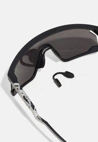 Schwarze Sonnenbrille mit verlaufenden dunklen Gläsern, ausgestattet mit einem strukturierten Metallrahmen, gebogenen Bügeln und abnehmbaren Ohrhaken.