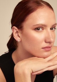 Boucles d'oreilles en argent cloutées avec un design rond, portées par une personne à la peau claire et aux longs cheveux brun-roux, le menton reposant sur les mains.