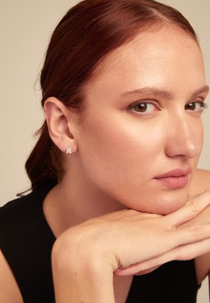 Boucles d'oreilles en argent cloutées avec un design rond, portées par une personne à la peau claire et aux longs cheveux brun-roux, le menton reposant sur les mains.