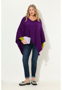 Poncho viola con scollo a V e spacchi laterali, indossato sopra una maglietta verde. Abbinato a jeans scuri e scarpe slip-on grigie, mentre tiene una borsa trapuntata lavanda.