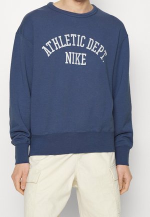 Osoba nosząca granatową bluzę z białym napisem "ATHLETIC DEPT. NIKE", w połączeniu z jasnobeżowymi spodnie cargo.
