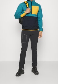 Pullover Columbia a mezza zip in teal e giallo, abbinato a pantaloni neri con tasca laterale e dettagli grigi. Scarpe nere completano l'outfit.