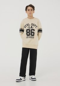 Felpa beige con lettering nero e righe, con la scritta "ATHL. DEPT. LA 86". I pantaloni neri e le sneakers bianche completano l'outfit.