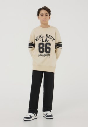 Felpa beige con lettering nero e righe, con la scritta "ATHL. DEPT. LA 86". I pantaloni neri e le sneakers bianche completano l'outfit.