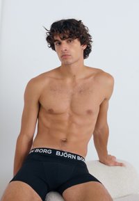 Crne bokserice s bijelim elastičnim pojasom na kojem piše "BJÖRN BORG". Model ima kovrčavu kosu i goli gornji dio tijela, sjedi na svijetloj pozadini.