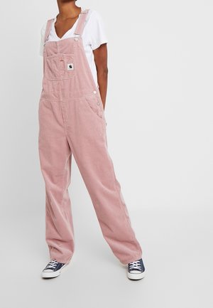 Vaaleanpunaiset, löysät corduroy-haalarit, joissa on etutasku, säädettävät olkaimet ja nepparikiinnitys. Yhdistetty valkoiseen t-paitaan ja tummansinisiin lenkkareihin.