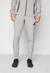 Hombre vestido con pantalones deportivos gris claro y chaqueta a juego con detalles de cremallera negra, y zapatillas deportivas negras, de pie sobre un suelo gris.