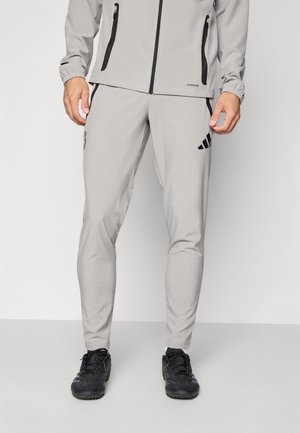 Uomo che indossa pantaloni atletici grigio chiaro e una giacca abbinata con dettagli in zip nera e scarpe sportive nere, in piedi su un pavimento grigio.