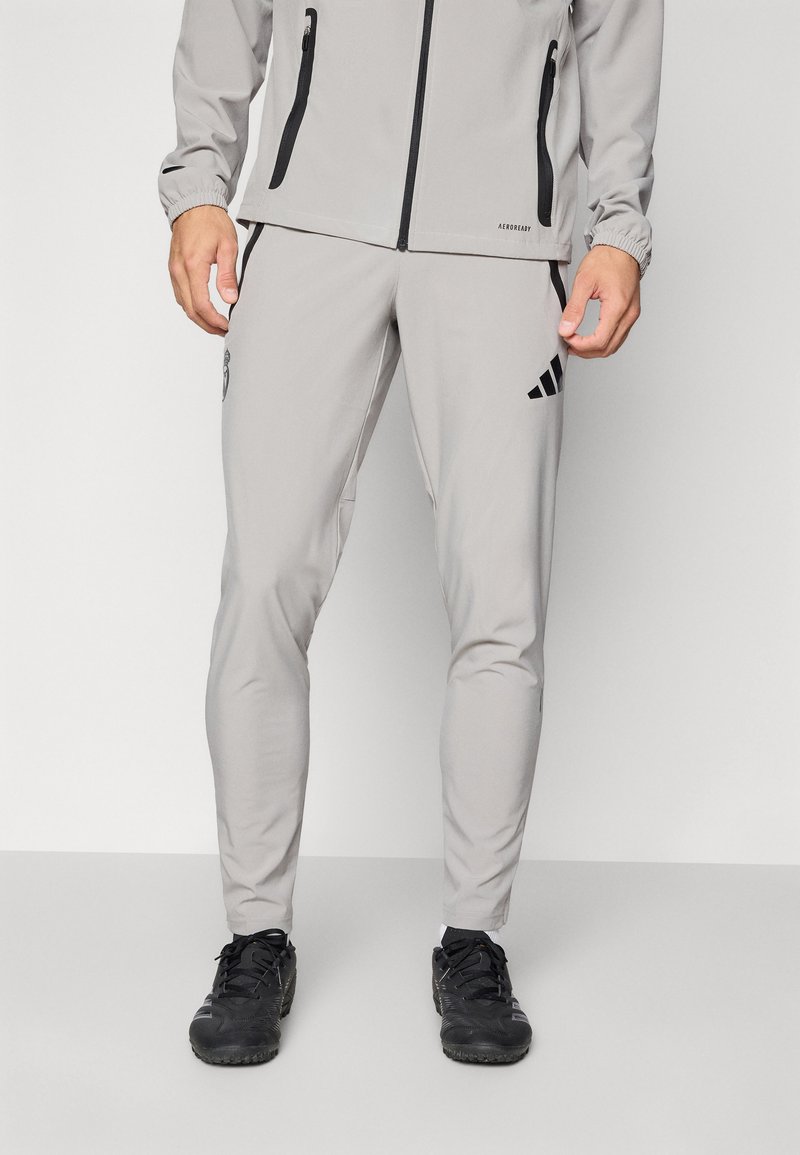 Hombre vestido con pantalones deportivos gris claro y chaqueta a juego con detalles de cremallera negra, y zapatillas deportivas negras, de pie sobre un suelo gris.