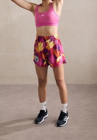 Rosa sport-BH, livfulla blommiga shorts med en logotyp, och svarta träningsskor med vita detaljer. Modell som står med armarna lyfta.