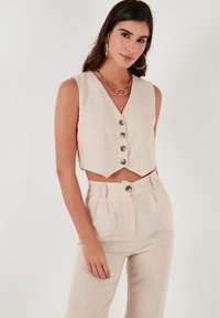 Liina crop-vest tumes beežis, millel on V-väljak, viis suurt nuppu ja laineline äär; koos sobivate kõrgete vöökohtade pükstega.
