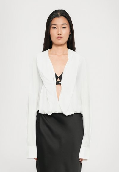 Femme aux longs cheveux foncés portant un chemisier blanc à col profond en V sur un soutien-gorge noir et une jupe noire en satin, debout devant un fond uni.