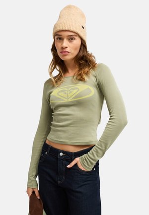 Jeune femme portant un bonnet en tricot beige, un crop top vert clair à manches longues avec un logo et un jean bleu foncé, posant avec une main dans la poche.
