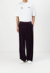 Vit bomulls-T-shirt med grafisk tryck och korta ärmar, i kombination med löst sittande, mörklila sammetbyxor och vita sneakers.