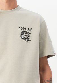 Světle šedé bavlněné tričko s černým vyšívaným logem "REPLAY" a designem lebky na levém prsu. Krátké rukávy, klasický střih.