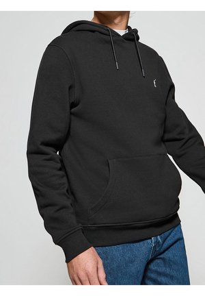 Mies pukeutuneena mustaan hupulliseen collegepaitaan, jossa on etutasku ja kiristysnyörit, yhdistettynä sinisiin farkkuihin, vaaleaa taustaa vasten.