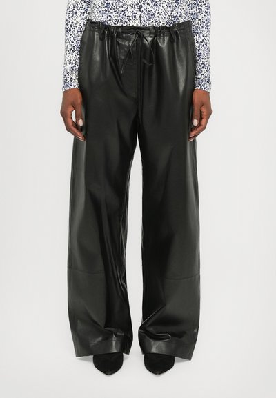 Pantalones de cuero negros de corte amplio, con cintura ajustable y textura suave. Combinados con zapatos negros de punta.