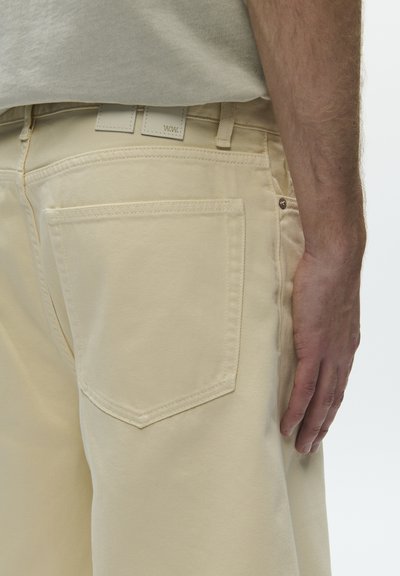 Zbliżenie osoby noszącej beżowe spodnie, ukazujące szczegóły tylnej kieszeni oraz część ramienia swobodnie opuszczonego wzdłuż ciała.