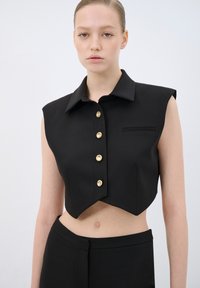 Maglietta nera cropped con colletto a punta, cinque bottoni dorati e un'unica tasca sul petto; realizzata in tessuto liscio con una vestibilità sartoriale.