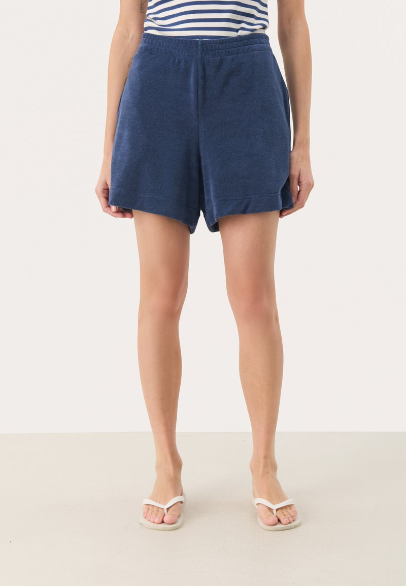 Shorts in morbido tessuto di spugna blu navy con vita elasticizzata, caratterizzati da una vestibilità leggermente ampia e orlo arrotolato. Indossati con sandali infradito bianchi.