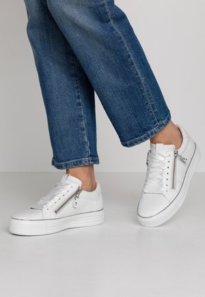 Ben i blå jeans iført hvide platformsneakers med sidelommer med lynlås og snørebånd, stående på en lysegrå gulv og baggrund.