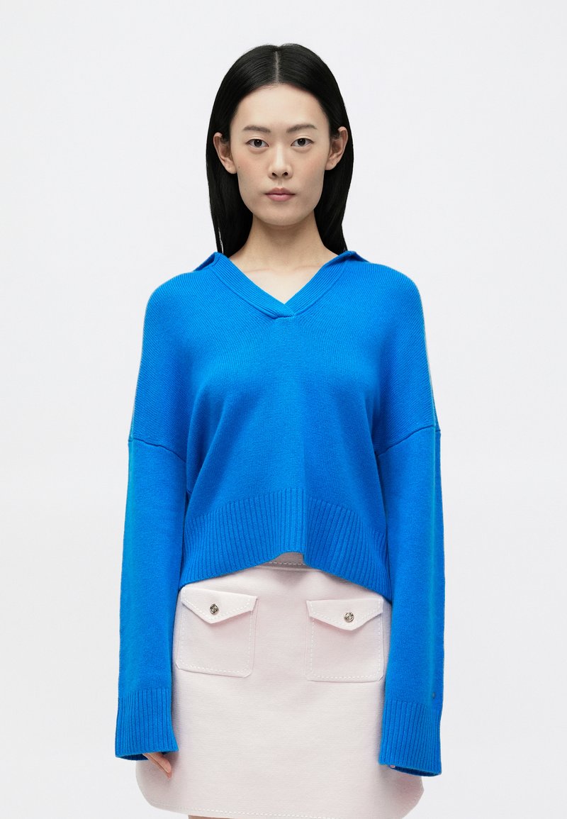 Niebieski sweter z dzianiny z dekoltem w serek i długimi rękawami, z prążkowanymi mankietami i dołem. Połączony z jasnoróżową spódnicą z detalami kieszeni.