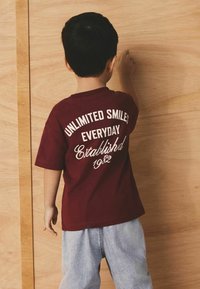 Temno rdeča majica s kratkimi rokavi s belim napisom "NEOMEJENI NASMEHI VSAK DAN Ustanovljeno 1982", kombinirana s svetlo modrimi denim hlačami.