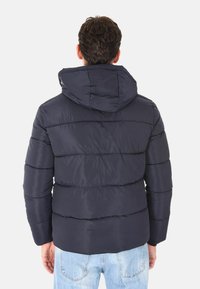Giacca piumino blu navy con cappuccio, caratterizzata da cuciture a sacchetto e una texture liscia. Abbinata a jeans in denim blu chiaro.