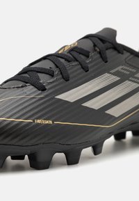 Black soccer cleat with a textured fiberskin upper, featuring silver stripes and gold accents, hexagonal studs, and black laces.