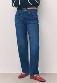 Tumši zili džinsi ar augstu jostasvietu un taisnām kājām, piecu kabatu un rāvējslēdzēja aizdare, kopā ar spīdīgām sarkankrāsām baleta kurpēm.