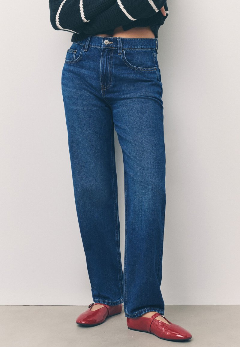 Tumši zili džinsi ar augstu jostasvietu un taisnām kājām, piecu kabatu un rāvējslēdzēja aizdare, kopā ar spīdīgām sarkankrāsām baleta kurpēm.