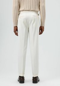 Witte op maat gemaakte broek met een gladde afwerking, voorzien van een rechte snit, plooien en geknoopte achterzakken. Gecombineerd met bruine schoenen.