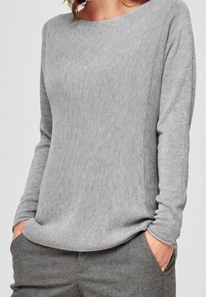 Woman wearing a light grey long-sleeve knit sweater with hands in pockets and matching grey pants, against a plain background.