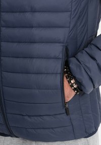 Granatowa kurtka puchowa z poziomymi przeszyciami, boczną kieszenią na zamek błyskawiczny i elastycznymi mankietami. Posiada teksturowane wykończenie i gładki materiał.