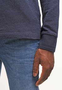 Long-sleeve navy shirt with a textured pattern, paired with blue denim jeans. Close-up of sleeve and hand on the thigh.