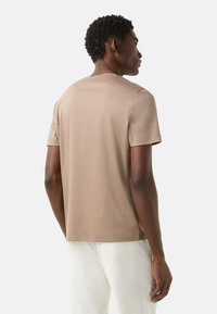 Koszulka z krótkim rękawem i okrągłym dekoltem w beżowym kolorze, wykonana z bawełny o gładkiej fakturze, z luźnym krojem, widziana od tyłu na jednolitym tle.